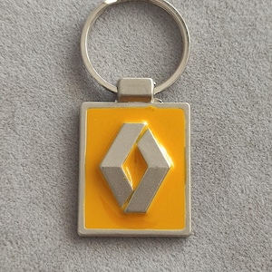 May include: A rectangular metal keychain featuring a bright yellow enamel background with a raised silver-toned Renault diamond logo in the center. The keychain includes a circular silver metal split ring attached to the top of the rectangular piece.