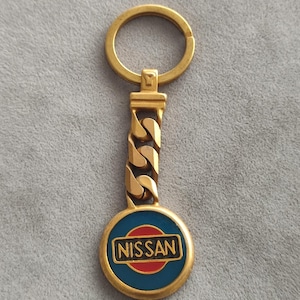 Peut inclure: Un porte-clés en métal doré composé d'un emblème circulaire avec une bordure bleue, un centre rouge et une barre rectangulaire noire affichant le mot NISSAN en lettres dorées. Le pendentif est relié à une courte chaîne et un anneau brisé.