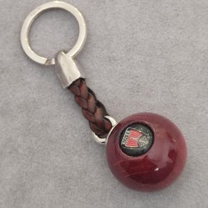 Può includere: Un portachiavi con un anello in metallo argentato fissato a un cinturino in pelle intrecciata marrone scuro, collegato a una sfera di legno liscia, rotonda e di colore rosso scuro. La sfera presenta un inserto nero con la scritta ROVER e un logo di nave vichinga rossa.