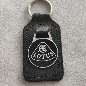 Könnte beinhalten: Ein schwarzer Schlüsselanhänger aus Leder mit einem rechteckigen Anhänger mit genähten Kanten und einem silbernen Metallring. Am Leder ist ein kreisförmiges schwarz-silbernes Emblem befestigt, das das Lotus-Logo mit dem Text LOTUS zeigt.