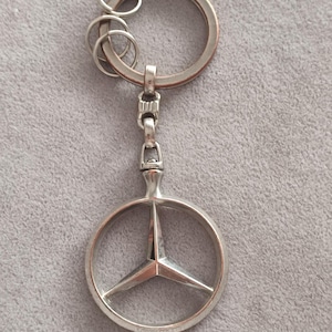 May include: A silver-toned metal keychain featuring the Mercedes-Benz three-pointed star logo enclosed within a circular frame. The keychain includes a large split ring and two smaller rings attached by a vertical metal link connector.