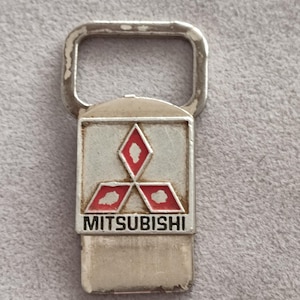 Könnte beinhalten: Ein silberfarbener Flaschenöffner aus Metall mit einem rechteckigen Emblem, das das Mitsubishi-Logo zeigt: drei rote Rauten in Dreiecksform auf weißem Grund, darunter der Schriftzug MITSUBISHI in schwarzen Großbuchstaben.