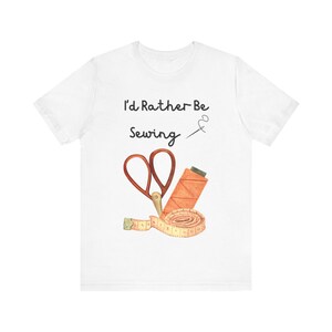May include: A white short-sleeve t-shirt featuring the black text I'd Rather Be Sewing, a small needle and thread icon, a pair of red-handled scissors, an orange spool of thread, and a coiled yellow measuring tape with visible markings.