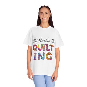 Peut inclure: Un t-shirt blanc à manches courtes avec le texte noir I'd Rather Be et le mot QUILTING en grandes lettres multicolores aux motifs complexes et vibrants. Le t-shirt est associé à un jean bleu clair, créant une tenue décontractée.