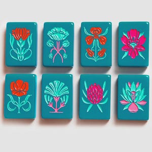 K&ouml;nnte beinhalten: Acht rechteckige, blaugr&uuml;ne Mahjong-Spielsteine, angeordnet in zwei Reihen zu je vier St&uuml;ck. Jeder Spielstein zeigt ein einzigartiges, erhabenes Blumenmuster in leuchtenden Farben wie Rot, Pink und Hellgr&uuml;n. Die Steine haben eine glatte, gl&auml;nzende Oberfl&auml;che.
