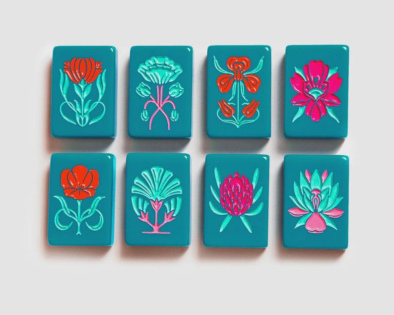K&ouml;nnte beinhalten: Acht rechteckige, blaugr&uuml;ne Mahjong-Spielsteine, angeordnet in zwei Reihen zu je vier St&uuml;ck. Jeder Spielstein zeigt ein einzigartiges, erhabenes Blumenmuster in leuchtenden Farben wie Rot, Pink und Hellgr&uuml;n. Die Steine haben eine glatte, gl&auml;nzende Oberfl&auml;che.