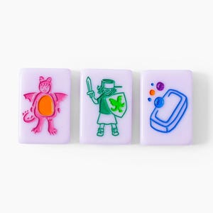 Pu&ograve; includere: Tre piastrelle rettangolari color viola chiaro con disegni colorati in rilievo. La prima piastrella mostra un drago rosa con la pancia arancione. La seconda raffigura una figura con una spada e uno scudo verde. La terza mostra una saponetta blu.