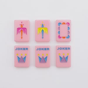 May include: Six rectangular pink mahjong tiles arranged in two rows of three. The top row features two tiles with stylized arrow designs and one with a circular border. The bottom row contains three identical tiles labeled JOKER with a crown graphic.