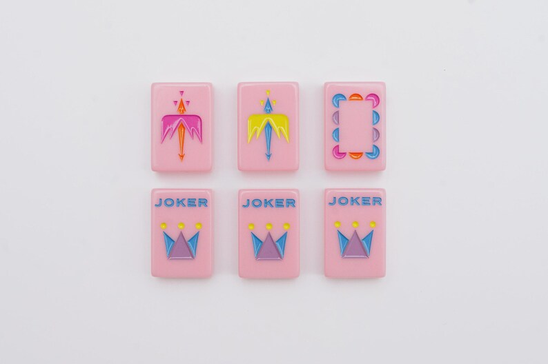 May include: Six rectangular pink mahjong tiles arranged in two rows of three. The top row features two tiles with stylized arrow designs and one with a circular border. The bottom row contains three identical tiles labeled JOKER with a crown graphic.