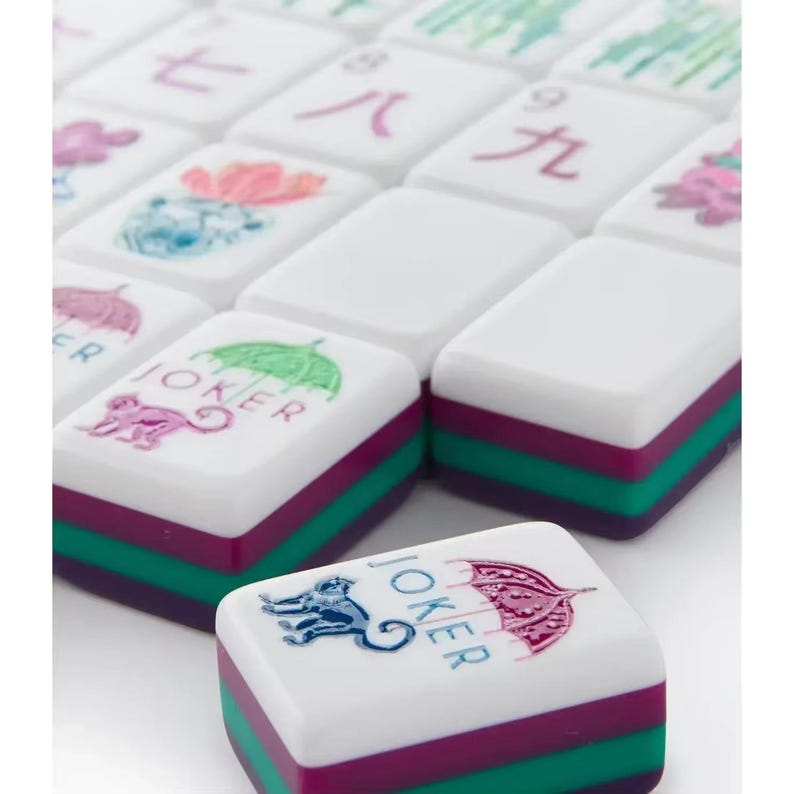 Może przedstawiać: Kolekcja białych płytek do gry w Mahjong z wielokolorowymi grafikami przedstawiającymi chińskie znaki, małpy i parasole. Każda płytka ma warstwową konstrukcję z białą g&oacute;rą, turkusową środkową sekcją i ciemnofioletową podstawą.