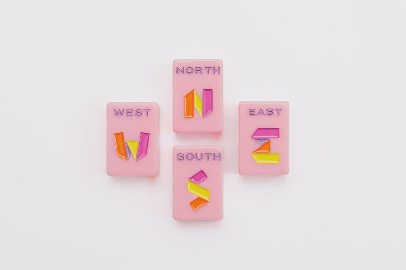 May include: Four rectangular pink tiles arranged in a compass formation. Each tile features a purple cardinal direction label&mdash;NORTH, SOUTH, EAST, or WEST&mdash;and a stylized, multi-colored letter representing that direction in orange, yellow, and magenta.
