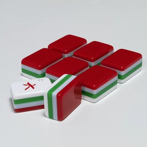 May include: A collection of rectangular mahjong tiles featuring a layered design with a red top, a green middle stripe, and a white base. One tile is positioned horizontally showing a red character symbol on its white face, while another stands vertically.