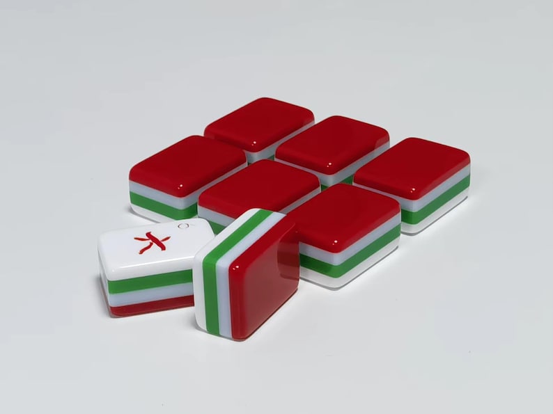 May include: A collection of rectangular mahjong tiles featuring a layered design with a red top, a green middle stripe, and a white base. One tile is positioned horizontally showing a red character symbol on its white face, while another stands vertically.