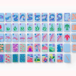 Puede incluir: Un conjunto de fichas de mahjong rectangulares de color azul claro con ilustraciones coloridas que incluyen notas musicales, plantas del desierto, rutas de mapas, patrones florales e iconos culturales. Algunas fichas muestran los n&uacute;meros del uno al nueve, otras las letras N, E, W, S o la palabra JOKER.