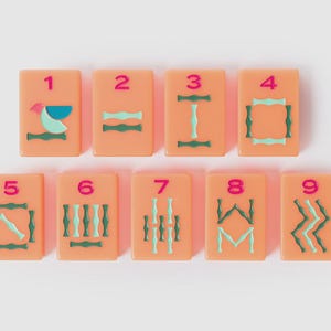 May include: A set of nine rectangular peach-colored mahjong tiles arranged in two rows. Each tile features a pink numeral from one to nine at the top, with various geometric and stylized green and light blue patterns engraved below the numbers.