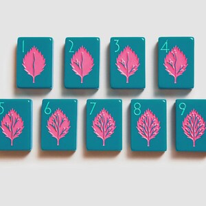 K&ouml;nnte beinhalten: Ein Set aus neun rechteckigen, blaugr&uuml;nen Mahjong-Spielsteinen, die in zwei Reihen angeordnet sind. Jeder Spielstein zeigt eine leuchtend rosa Blattillustration in der Mitte sowie eine hellblaue Ziffer von eins bis neun in der oberen linken Ecke.