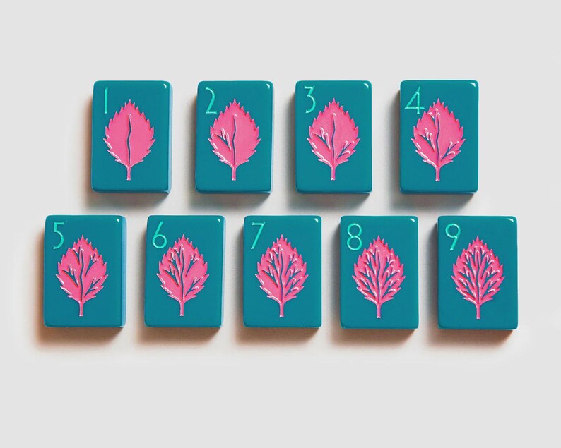 K&ouml;nnte beinhalten: Ein Set aus neun rechteckigen, blaugr&uuml;nen Mahjong-Spielsteinen, die in zwei Reihen angeordnet sind. Jeder Spielstein zeigt eine leuchtend rosa Blattillustration in der Mitte sowie eine hellblaue Ziffer von eins bis neun in der oberen linken Ecke.