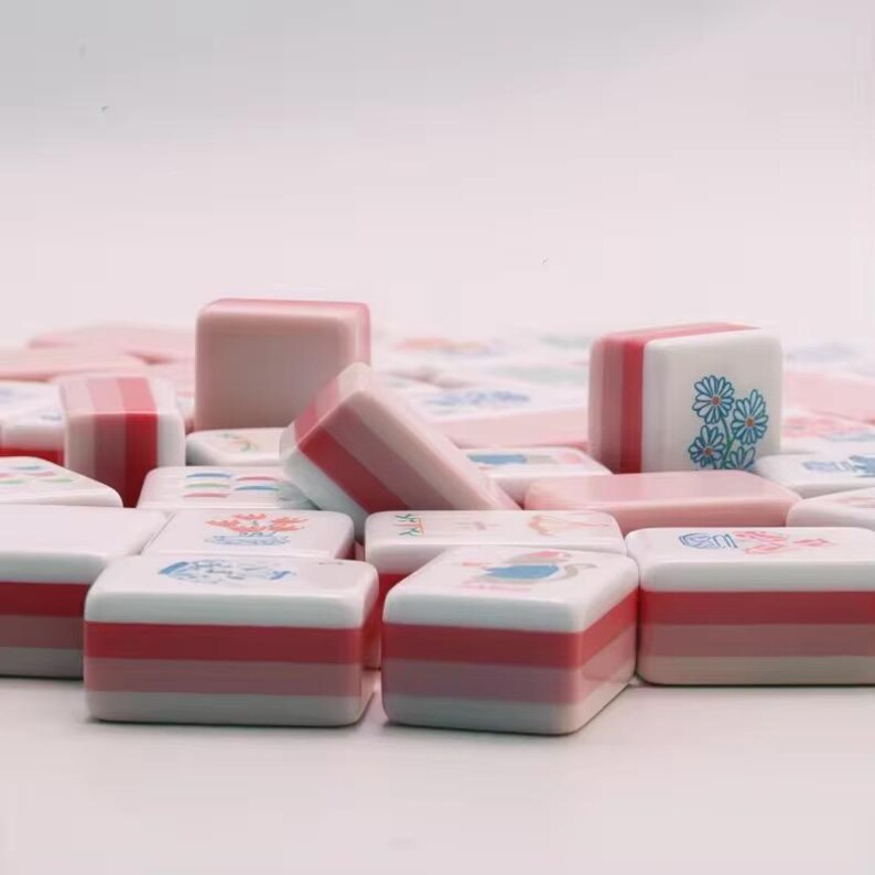 May include: A collection of rectangular mahjong tiles featuring a layered pink and white color scheme. Each tile displays a unique graphic illustration on its top surface, including floral patterns, small animals, and various decorative symbols.