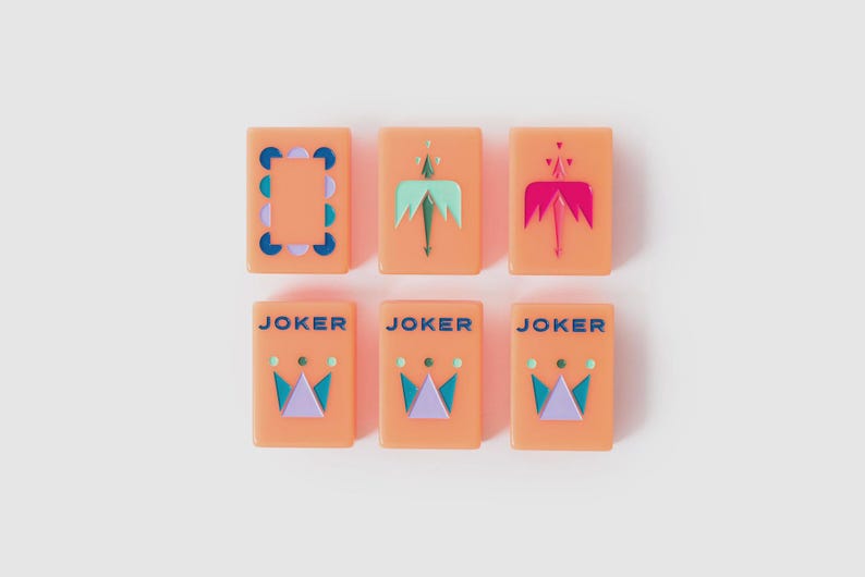 May include: Six rectangular peach-colored tiles arranged in two rows. The top row features a patterned border tile, a light green arrow design, and a dark pink arrow design. The bottom row contains three identical tiles labeled JOKER in blue text above a crown graphic in light green, dark blue, and light purple.