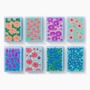 Puede incluir: Ocho azulejos rectangulares con bordes azul claro, cada uno con un patr&oacute;n floral en relieve &uacute;nico en colores vibrantes que incluyen rosa, naranja, morado y azul, dispuestos en dos filas de cuatro sobre un fondo blanco liso.