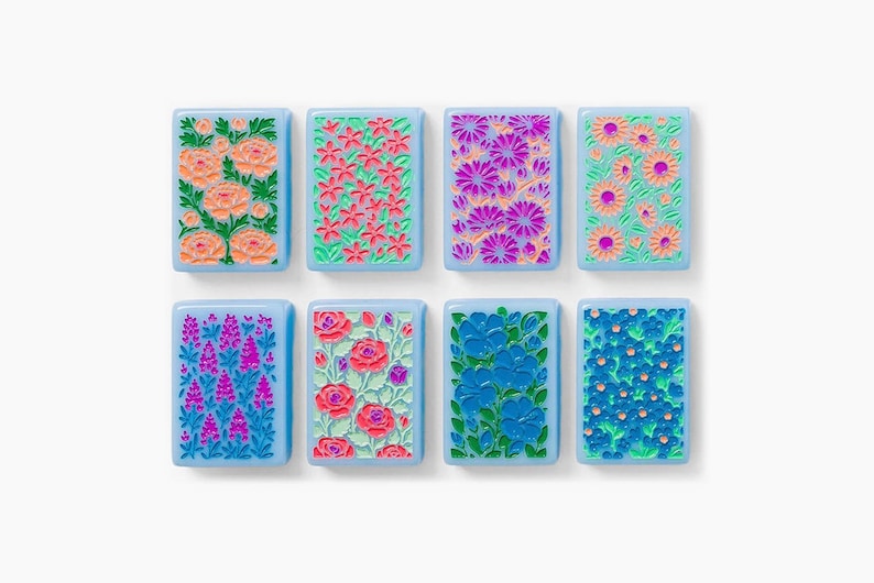 Puede incluir: Ocho azulejos rectangulares con bordes azul claro, cada uno con un patr&oacute;n floral en relieve &uacute;nico en colores vibrantes que incluyen rosa, naranja, morado y azul, dispuestos en dos filas de cuatro sobre un fondo blanco liso.
