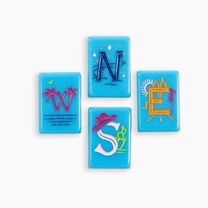 May include: Four rectangular light blue tiles arranged in a compass formation. Each tile features a letter: W with pink palm trees, N with dark blue waves and stars, E with an orange sun and city skyline, and S with a pink cowboy hat and cactus.