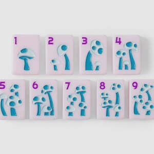 Puede incluir: Un conjunto de nueve baldosas rectangulares de color rosa claro, cada una con un n&uacute;mero morado del uno al nueve y un dise&ntilde;o de hongo azul en relieve. El n&uacute;mero de hongos en cada baldosa corresponde al d&iacute;gito morado en la esquina superior.