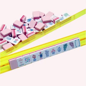 May include: A set of Mahjong tiles with pink backs and white faces featuring various colorful illustrations, including a monkey, flowers, teapots, and dragons, arranged on two translucent yellow plastic racks with a pile of extra tiles.