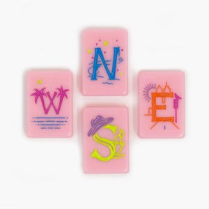 May include: Four rectangular pink tiles arranged in a cross shape, each featuring a letter representing a cardinal direction. The tiles display the letters W, N, E, and S, decorated with colorful graphic elements including palm trees, stars, a sun, and a hat.