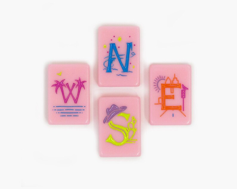 May include: Four rectangular pink tiles arranged in a cross shape, each featuring a letter representing a cardinal direction. The tiles display the letters W, N, E, and S, decorated with colorful graphic elements including palm trees, stars, a sun, and a hat.