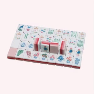 May include: A set of white mahjong tiles with pink backs, featuring colorful illustrations of teapots, teacups, flowers, dragons, and monkeys. Some tiles are standing upright, while others are laid flat in a grid, displaying various numbers and symbols.