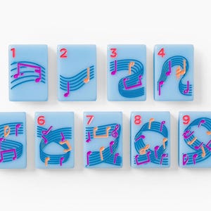 Puede incluir: Un conjunto de nueve fichas rectangulares de color azul claro, cada una con un n&uacute;mero rojo del uno al nueve en la esquina superior izquierda. Las fichas est&aacute;n decoradas con pentagramas azul oscuro y notas musicales rosas y naranjas.