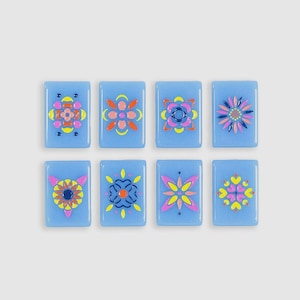 May include: Eight rectangular light blue glass tiles arranged in two rows of four. Each tile features a unique, symmetrical floral or geometric pattern painted in vibrant colors including pink, yellow, orange, and dark blue, creating a set.
