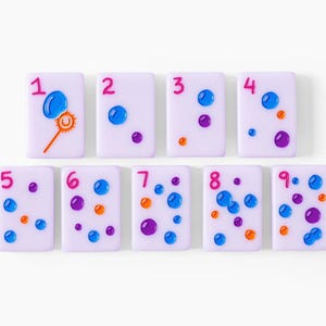 Pu&ograve; includere: Nove tessere rettangolari color viola chiaro disposte su due file, ognuna con un numero rosa da uno a nove e dettagli in rilievo lucidi blu, viola e arancioni. La prima tessera mostra l'icona di una bacchetta per fare le bolle di sapone.
