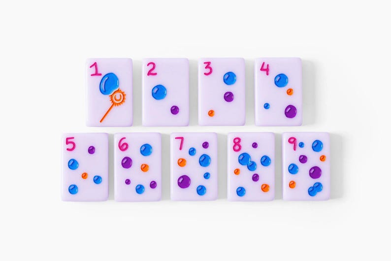 Pu&ograve; includere: Nove tessere rettangolari color viola chiaro disposte su due file, ognuna con un numero rosa da uno a nove e dettagli in rilievo lucidi blu, viola e arancioni. La prima tessera mostra l'icona di una bacchetta per fare le bolle di sapone.