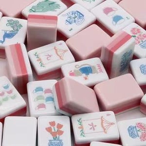 May include: A collection of rectangular mahjong tiles in shades of pink and white. The tiles feature various illustrations including a green dragon, a blue teapot, pink tulips, a monkey, and tea cups, alongside Chinese characters and the word Joker.