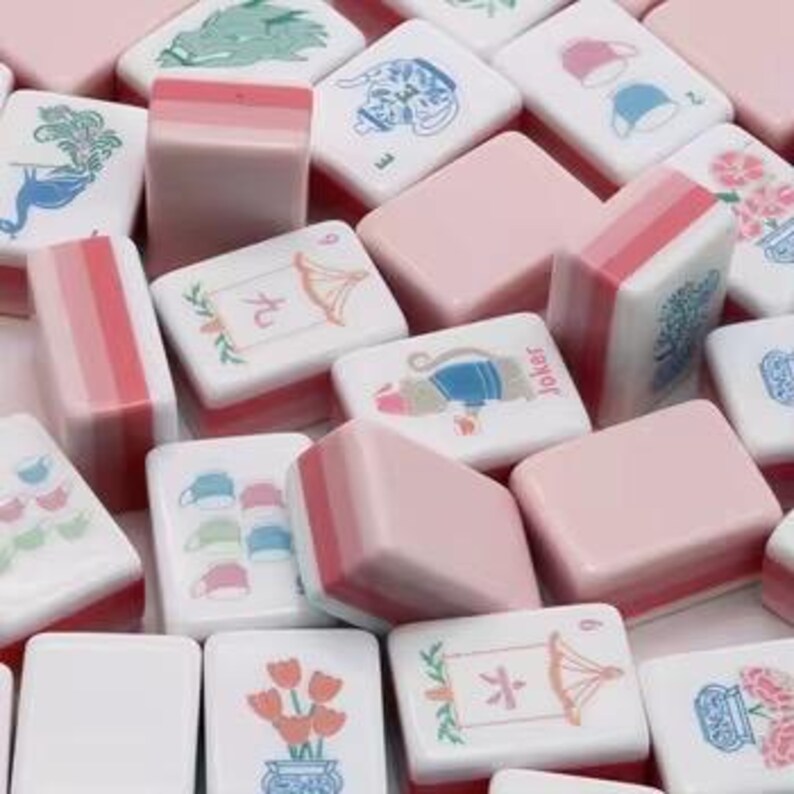 May include: A collection of rectangular mahjong tiles in shades of pink and white. The tiles feature various illustrations including a green dragon, a blue teapot, pink tulips, a monkey, and tea cups, alongside Chinese characters and the word Joker.