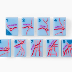 Puede incluir: Un conjunto de nueve azulejos rectangulares de color azul claro, cada uno con un dise&ntilde;o de mapa de carreteras en relieve rojo, una etiqueta numerada del uno al nueve y un nombre de ciudad espec&iacute;fico, incluyendo Los Angeles, Flagstaff, Santa Fe, Amarillo, Tulsa, Joplin, St. Louis, Springfield y Chicago.