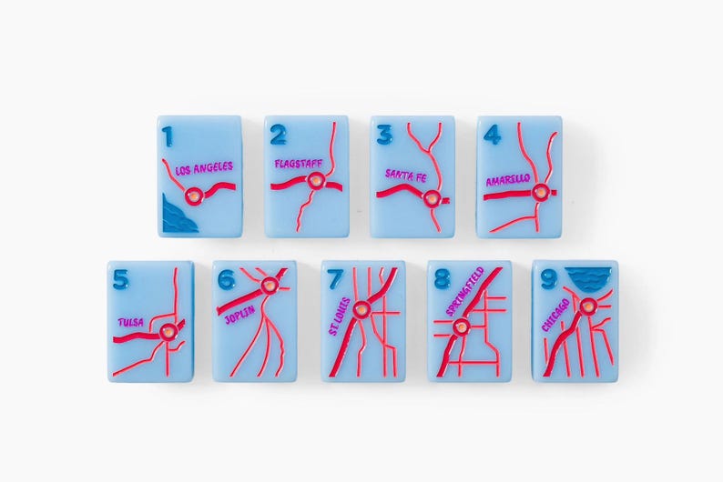 Puede incluir: Un conjunto de nueve azulejos rectangulares de color azul claro, cada uno con un dise&ntilde;o de mapa de carreteras en relieve rojo, una etiqueta numerada del uno al nueve y un nombre de ciudad espec&iacute;fico, incluyendo Los Angeles, Flagstaff, Santa Fe, Amarillo, Tulsa, Joplin, St. Louis, Springfield y Chicago.