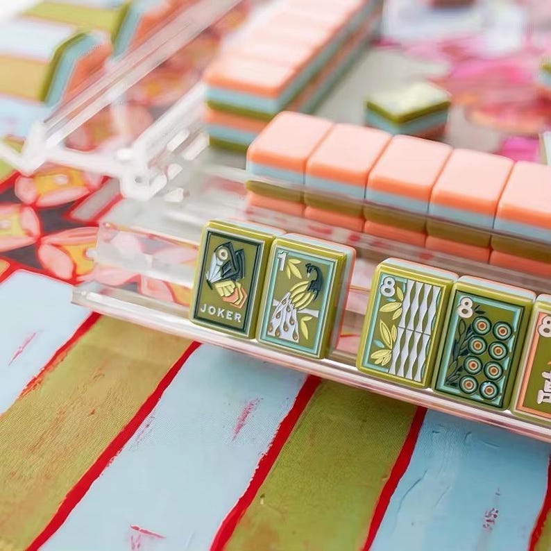 May include: A set of Mahjong tiles arranged on a clear acrylic rack. The tiles feature olive green faces with detailed illustrations of a Joker, a peacock, and numerical patterns. Behind the rack, stacked tiles show peach and light blue sides.