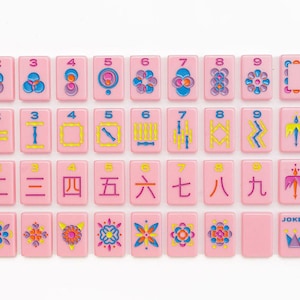 May include: A set of rectangular pink mahjong tiles arranged in four rows. The tiles feature colorful engraved designs including numbers one through nine, directional markers labeled North, East, South, West, and various floral and geometric patterns.