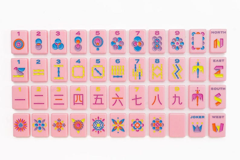 May include: A set of rectangular pink mahjong tiles arranged in four rows. The tiles feature colorful engraved designs including numbers one through nine, directional markers labeled North, East, South, West, and various floral and geometric patterns.