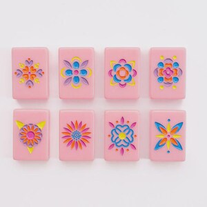 May include: Eight rectangular pink tiles arranged in two rows of four. Each tile features a unique, recessed floral or geometric pattern engraved into the surface, filled with vibrant colors including yellow, blue, orange, and magenta.