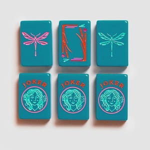 K&ouml;nnte beinhalten: Sechs rechteckige, blaugr&uuml;ne Mahjong-Spielsteine in zwei Reihen. Die obere Reihe zeigt zwei Steine mit rosa Libellen und einen mit einem roten Rahmen. Die untere Reihe zeigt drei JOKER-Steine mit einer kreisf&ouml;rmigen Grafik einer Person.