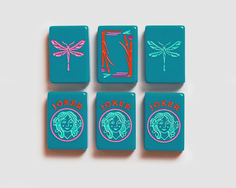 K&ouml;nnte beinhalten: Sechs rechteckige, blaugr&uuml;ne Mahjong-Spielsteine in zwei Reihen. Die obere Reihe zeigt zwei Steine mit rosa Libellen und einen mit einem roten Rahmen. Die untere Reihe zeigt drei JOKER-Steine mit einer kreisf&ouml;rmigen Grafik einer Person.