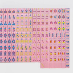 May include: A set of pink Mahjong tiles arranged in a grid, featuring colorful engraved numbers, symbols, and characters in blue, yellow, and red. The tiles include numbered suits, directional markers, floral patterns, and Joker tiles with crown icons.