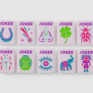 Puede incluir: Diez fichas rectangulares de color rosa claro, cada una con el texto JOKER en morado. Las fichas presentan iconos en relieve de colores que incluyen una herradura, una estrella fugaz, un Buda, un tr&eacute;bol, una mariquita, un escarabajo, un tarro, un ojo, un elefante y una figura estilizada.