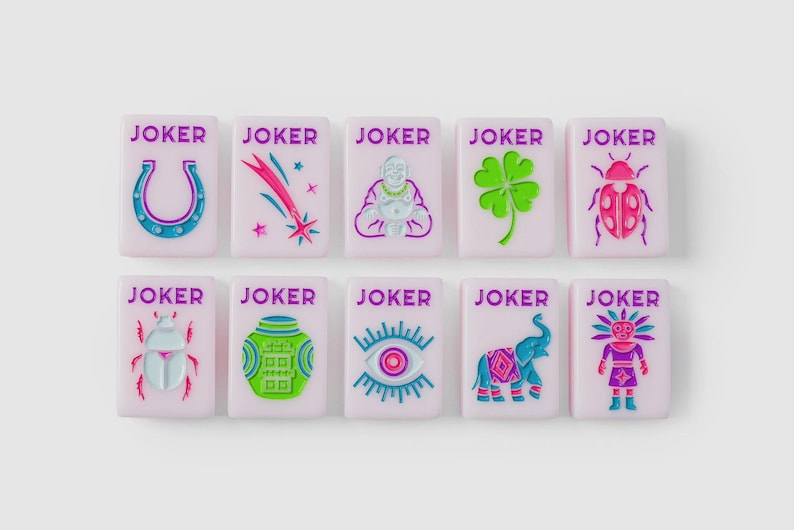 Puede incluir: Diez fichas rectangulares de color rosa claro, cada una con el texto JOKER en morado. Las fichas presentan iconos en relieve de colores que incluyen una herradura, una estrella fugaz, un Buda, un tr&eacute;bol, una mariquita, un escarabajo, un tarro, un ojo, un elefante y una figura estilizada.