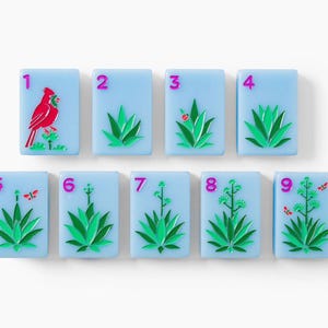 Puede incluir: Un conjunto de nueve fichas de mahjong rectangulares de color azul claro dispuestas en dos filas. Cada ficha presenta un n&uacute;mero magenta del uno al nueve y una ilustraci&oacute;n de una planta de agave verde. La primera incluye un cardenal rojo.