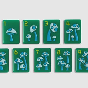 May include: A set of nine rectangular green tiles, each featuring a yellow number from one to nine and a raised blue and light blue mushroom design. The number of mushrooms on each tile corresponds to the numerical value displayed on the tile.